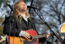 Sean Feucht speaking at a large outdoor worship rally with flags and supporters in the background