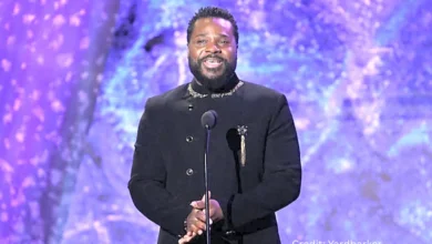 Malcolm‑Jamal Warner speaking at a creative arts showcase, wearing a black shirt and holding a microphone